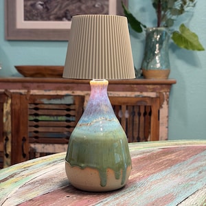 May include: A table lamp with a beige, vertically ribbed, conical lampshade and a ceramic base. The base features a teardrop shape with a glaze that transitions from a light purple at the neck to a vibrant blue and a dripping sage green finish.