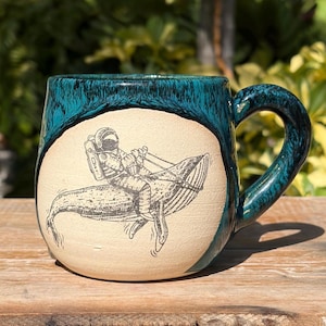 May include: A ceramic mug with a glossy teal drip glaze over a matte cream-colored base. The front features a detailed black line drawing of an astronaut riding on the back of a whale, with the astronaut holding onto reins attached to the whale.