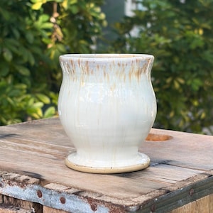 May include: A ceramic mug with a rounded body and a flared rim, featuring a glossy off-white glaze with vertical tan and brown drip patterns cascading from the top edge down the sides, resting on a circular base with a smooth, light tan finish.