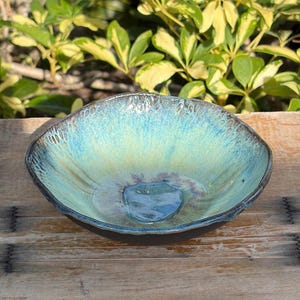 May include: A shallow ceramic bowl with a reactive glaze featuring a gradient of turquoise, teal, and pale yellow. The rim has a textured, dark brown edge, and the interior surface shows a smooth, glossy finish with a deep blue center pool.