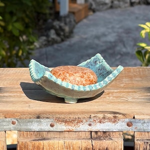 May include: A round, textured orange soap bar rests inside a shallow, hand-formed ceramic dish. The dish features a scalloped, ruffled edge with a glossy turquoise and teal glaze, showcasing a mottled, variegated color pattern throughout.