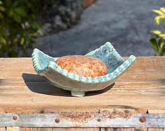 May include: A round, textured orange soap bar rests inside a shallow, hand-formed ceramic dish. The dish features a scalloped, ruffled edge with a glossy turquoise and teal glaze, showcasing a mottled, variegated color pattern throughout.