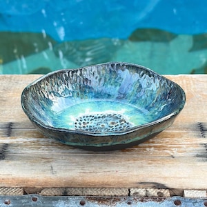 May include: A shallow, round ceramic bowl with an irregular, wavy rim. The interior features a glossy glaze transitioning from deep navy blue at the edges to a bright turquoise and light green center, accented by a dark, textured, dotted pattern.