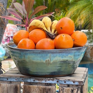 May include: A large, round ceramic bowl with a multi-toned blue and green glaze, filled with a cluster of fresh oranges and a bunch of bananas. The bowl features a smooth, glossy finish with visible color variations and a dark, earthy rim.
