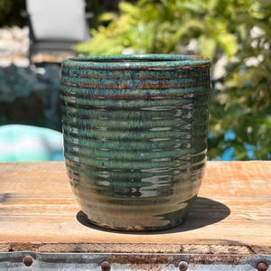 May include: A handmade ceramic tumbler with a textured, ridged surface. The glaze is a blend of greens and browns, creating a natural, earthy aesthetic. The cylindrical shape is ideal for beverages.