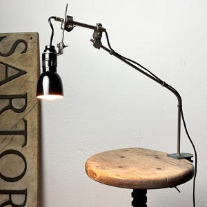 Può includere: Una lampada da tavolo in metallo nero con braccio articolato regolabile e base a morsetto. La lampada presenta un paralume nero lucido a forma di cupola, un cavo di alimentazione nero e giunti metallici. La luce è accesa e illumina verso il basso.