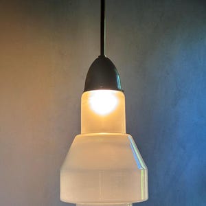 Op de afbeelding: Een vintage hanglamp met een matglazen kap en een donkere metalen steel. De gloeilamp is verlicht en geeft een warme gloed. De kap heeft een uniek, gelaagd ontwerp. De armatuur is opgehangen aan een plafondbevestiging.