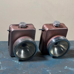 Könnte beinhalten: Zwei vintage-Taschenlampen aus braunem Metall mit silbernen Akzenten. Jede Taschenlampe hat eine runde, klare Glasscheibe und einen silbernen Knopf oben.