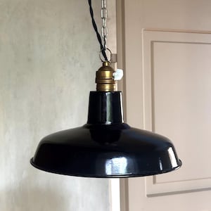 Op de afbeelding: Een zwarte hanglamp met een glanzende afwerking, voorzien van een messingkleurige fitting met een witte draaischakelaar. De lamp hangt aan een zilverkleurige metalen ketting en een zwart gedraaid stoffen snoer voor een industriële uitstraling.