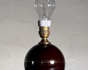 May include: A vintage table lamp featuring a dark red, spherical base with a gold band. It has a brass neck and a white socket, topped with a clear light bulb. The lamp's base is approximately 15 cm in diameter.