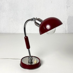 Könnte beinhalten: Eine Vintage-Schreibtischlampe mit bordeauxfarbenem Schirm und Sockel sowie einem verchromten, gebogenen Arm. Die Lampe hat einen kleinen, runden Sockel mit Schalter und einem bordeauxfarbenen Griff. Die Lampe ist für einen Schreibtisch oder Tisch konzipiert.