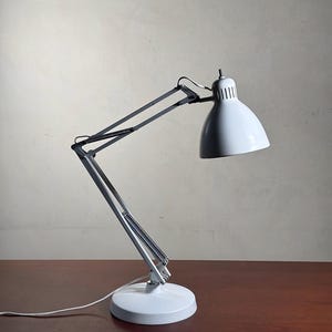 Puede incluir: Una lámpara de escritorio blanca con una pantalla curva y un brazo ajustable. La lámpara tiene una base redonda y un cable. La lámpara está sobre una superficie de madera.