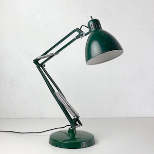 Puede incluir: Una lámpara de escritorio de metal verde oscuro con un brazo articulado ajustable con resortes plateados expuestos, una pantalla en forma de cúpula con interior blanco y una base circular. La lámpara incluye un cable negro.