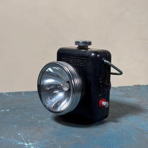 Op de afbeelding: Een zwarte Superbilt zaklamp met een zilveren reflector en een rode schakelaar. De zaklamp heeft een draagbeugel en een kleine knop bovenop.