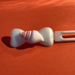 Puede incluir: Una pinza para el pelo de plástico blanco con un diseño en forma de lazo. El lazo presenta un acento en forma de cuerno a rayas rosas y blancas. La pinza está sobre un fondo naranja. Este accesorio para el cabello es adecuado para varios peinados.