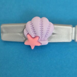 May include: A silver-coloured hair clip featuring a light purple seashell charm with vertical ridges and a small, solid pink starfish attached to the front. The accessory has a smooth, metallic finish and a simple, functional design for securing hair.