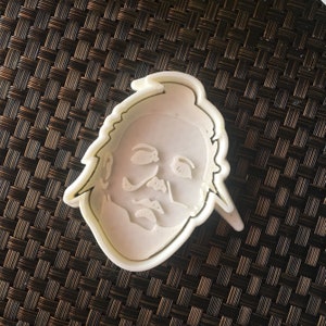 May include: White plastic cookie cutter shaped like a person's face with a serious expression.