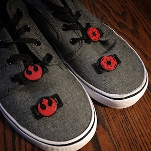 May include: A pair of gray canvas shoes with black laces and red Star Wars themed shoe charms. The charms are the Rebel Alliance symbol and the Imperial symbol.
