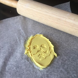 May include: A yellow plastic cookie cutter shaped like the face of a horror movie character with a white mask. The cookie cutter is on a piece of baking parchment next to a wooden rolling pin.