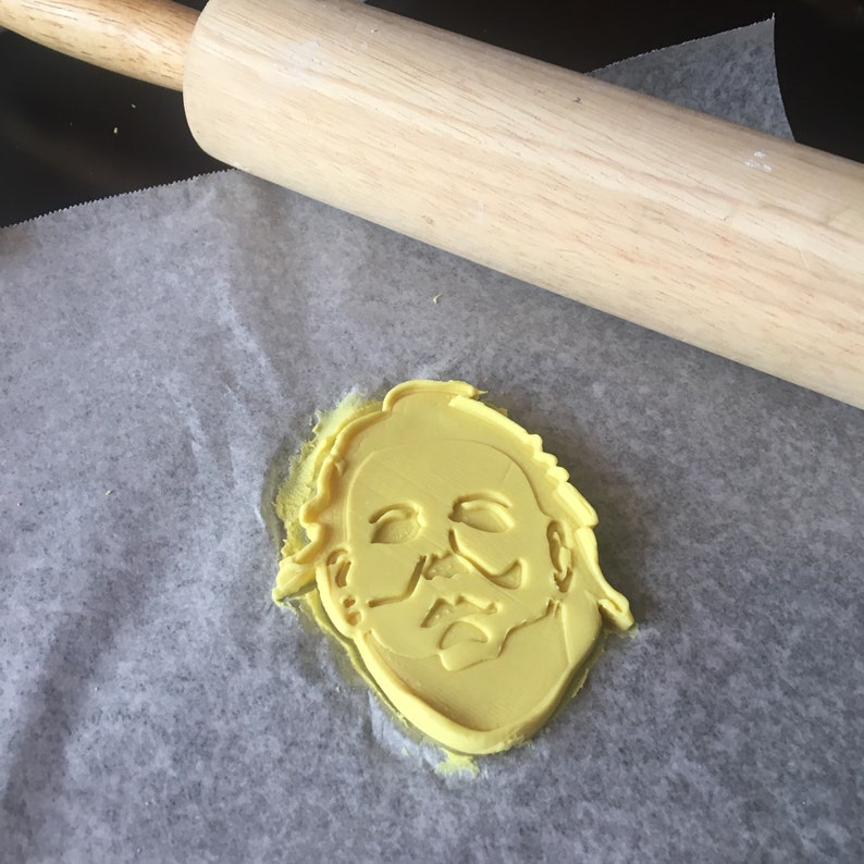 May include: A yellow plastic cookie cutter shaped like the face of a horror movie character with a white mask. The cookie cutter is on a piece of baking parchment next to a wooden rolling pin.