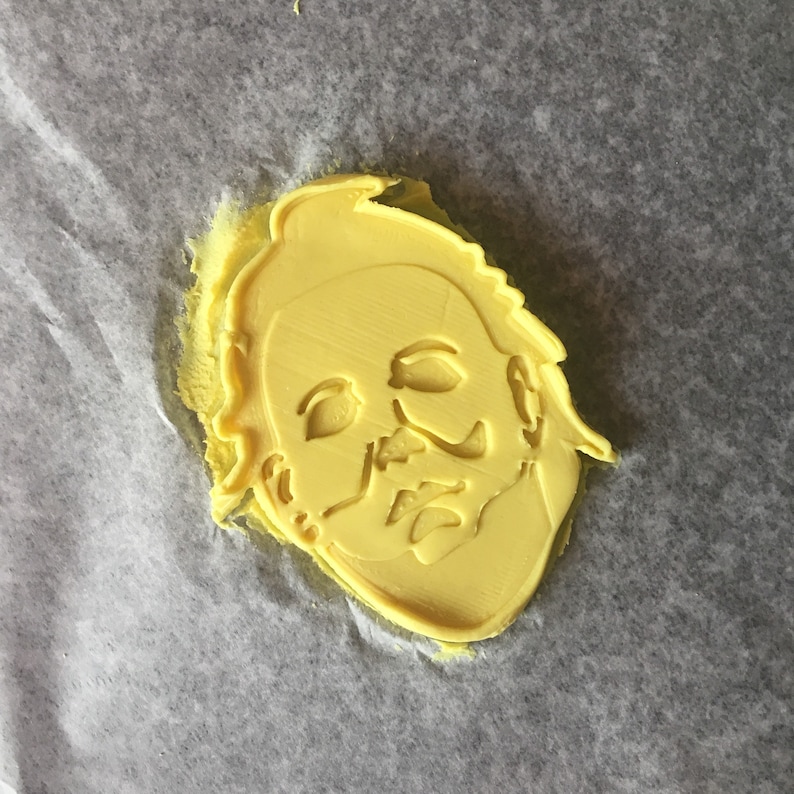 May include: Yellow plastic cookie cutter shaped like a person wearing a mask. The mask has a wide, flat shape with a prominent nose and eyes.