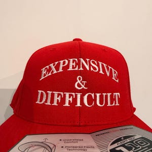 May include: A red baseball cap with white embroidered text reading "EXPENSIVE & DIFFICULT". The cap has a curved peak and a Flexfit Tech label. The hat is a one-size-fits-all OSFA cap.