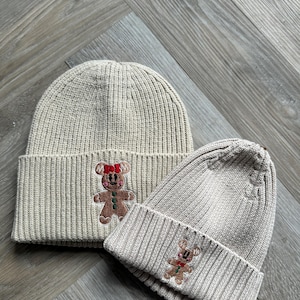 May include: Two beige knitted beanies with a ribbed design. Each beanie features an embroidered gingerbread man with a red bow and green accents. These beanies are a winter accessory, ideal for cold weather.