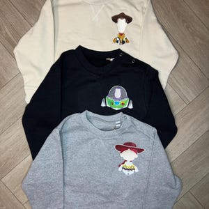 以下が含まれることがあります： トイストーリーのキャラクターのアップリケが付いた長袖スウェットシャツが3枚。1枚目はベージュでウッディ、2枚目は黒でバズ・ライトイヤー、3枚目はグレーでジェシーです。