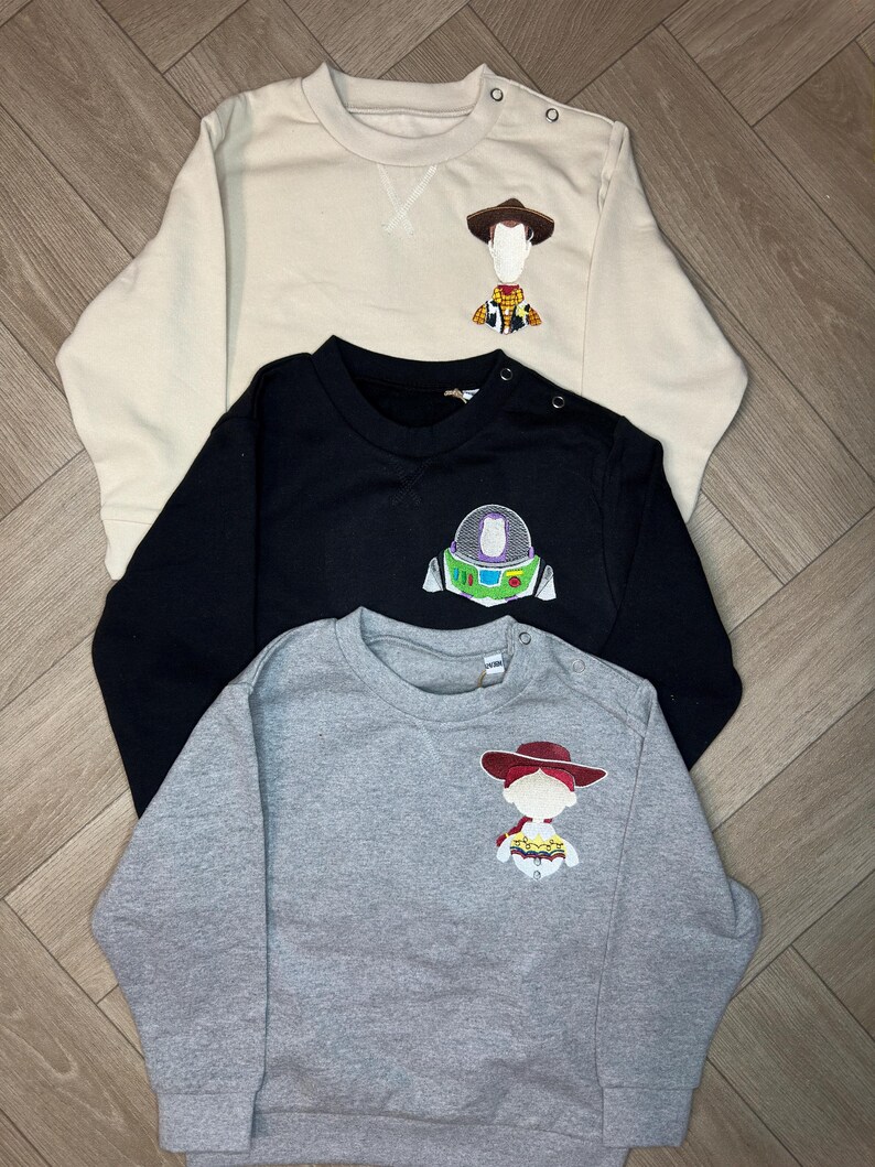 以下が含まれることがあります： トイストーリーのキャラクターのアップリケが付いた長袖スウェットシャツが3枚。1枚目はベージュでウッディ、2枚目は黒でバズ・ライトイヤー、3枚目はグレーでジェシーです。