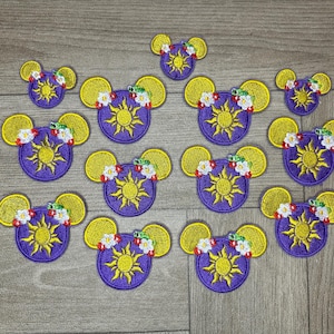 Op de afbeelding: Een set van 12 paarse en gele geborduurde strijkpatches met een cartoonzon met een gezicht en bloemen. De patches zijn gevormd als Mickey Mouse-oren.