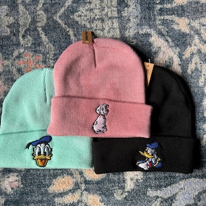 May include: Three beanies in mint green, pink, and black. Each beanie has a different embroidered cartoon character: Donald Duck, a Dalmatian puppy, and Donald Duck again. The beanies are folded at the brim.