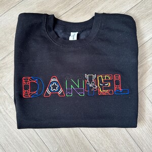 Puede incluir: Sudadera negra con el nombre "DANIEL" bordado en letras coloridas con tem&aacute;tica de superh&eacute;roes. Las letras est&aacute;n delineadas en varios colores, cada una con un dise&ntilde;o diferente, incluyendo una telara&ntilde;a, una estrella y un rayo.
