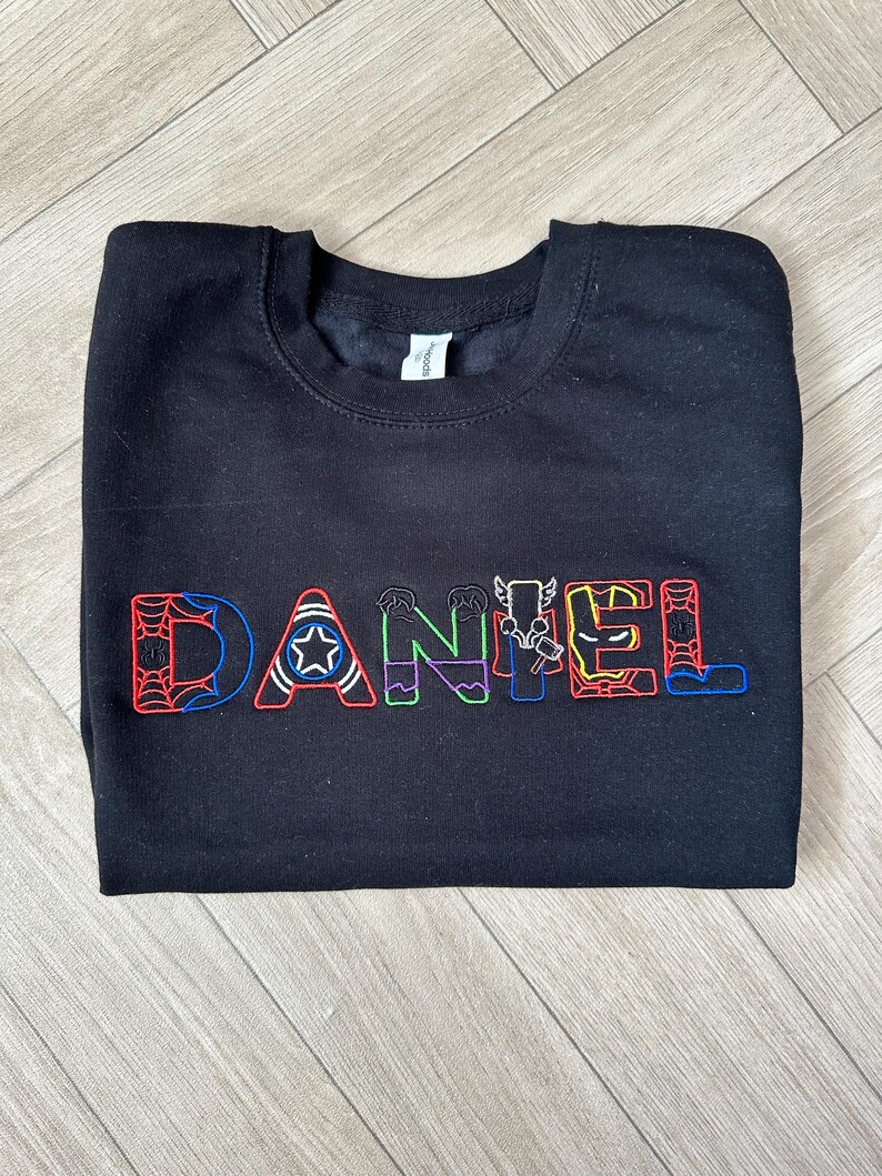 Puede incluir: Sudadera negra con el nombre "DANIEL" bordado en letras coloridas con tem&aacute;tica de superh&eacute;roes. Las letras est&aacute;n delineadas en varios colores, cada una con un dise&ntilde;o diferente, incluyendo una telara&ntilde;a, una estrella y un rayo.