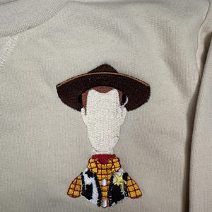 以下が含まれることがあります： ベージュのトレーナーに、トイストーリーのウッディの刺繍が施されています。ウッディは、トレードマークのテンガロンハット、ベスト、保安官のバッジを着用しています。