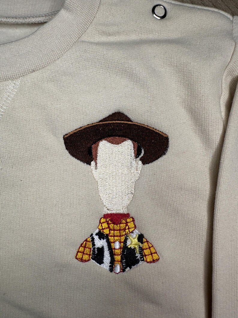 以下が含まれることがあります： ベージュのトレーナーに、トイストーリーのウッディの刺繍が施されています。ウッディは、トレードマークのテンガロンハット、ベスト、保安官のバッジを着用しています。