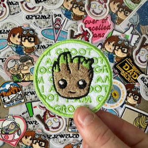 以下が含まれることがあります： ガーディアンズ・オブ・ギャラクシーのキャラクター、グルートが描かれた丸い刺繍パッチ。パッチは緑色の縁取りで、「I AM GROOT」の文字がキャラクターの周りにプリントされています。他のパッチが背景にあります。