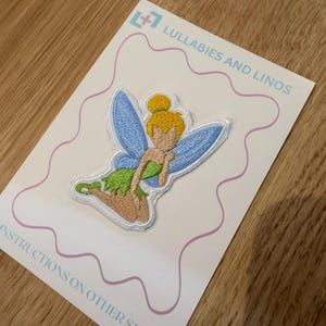 Può includere: Toppa ricamata raffigurante una fata con ali blu, un vestito verde e capelli biondi. La toppa è su una carta rosa chiaro con il testo "Lullabies and Linos" e "Instructions on other side".