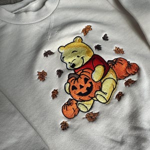 Può includere: Felpa per neonati color crema con ricamo del personaggio di Winnie the Pooh che tiene in mano una zucca di Halloween. Il design include zucche e foglie autunnali. La felpa ha uno scollo tondo e chiusure a scatto.