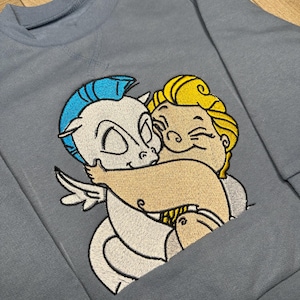 Könnte beinhalten: Hellblaues Baby-Sweatshirt mit einem gestickten Design von zwei Cartoon-Figuren, die sich umarmen. Eine Figur hat gelbes Haar, die andere blaues Haar. Das Sweatshirt hat einen Rundhalsausschnitt und Druckknöpfe an der Schulter.