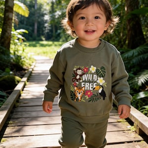 May include: A child in an olive green sweatshirt and trousers, smiling on a wooden bridge. The sweatshirt has a jungle-themed design with a monkey, tiger, toucan, and the words "WILD FREE". The background shows a green forest with trees.