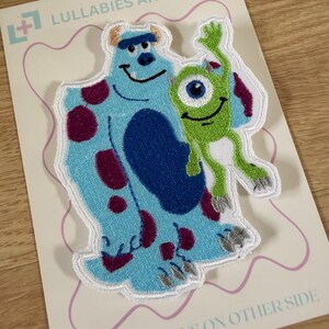 Peut inclure: Un écusson brodé représentant un grand monstre bleu à taches violettes et un plus petit monstre vert à un seul œil. L'écusson est fixé sur une carte blanche avec le texte LULLABIES AND LINOS en haut et INSTRUCTIONS ON OTHER SIDE en bas.