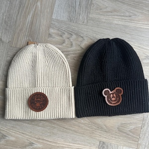May include: Two ribbed knit beanies. One is beige with a brown leather patch that says "OHANA COFFEE". The other is black with a brown leather Mickey Mouse patch. Both beanies have a folded brim.