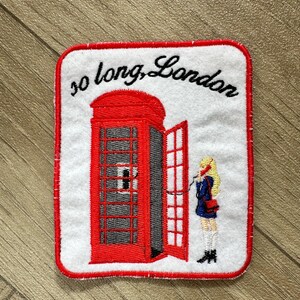 May include: Embroidered patch featuring a red London telephone booth with an open door. A blonde figure in a blue coat stands beside the booth. The text "so long, London" is arched above the booth.