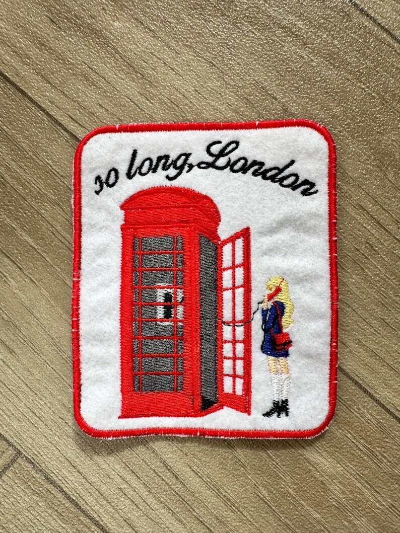 May include: Embroidered patch featuring a red London telephone booth with an open door. A blonde figure in a blue coat stands beside the booth. The text "so long, London" is arched above the booth.