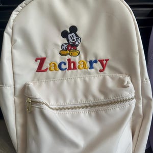 Peut inclure: Sac à dos crème avec une broderie de Mickey Mouse et le nom "Zachary" en lettres colorées. Il possède une poche avant zippée et une fermeture éclair dorée.