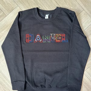 Puede incluir: Sudadera negra de cuello redondo con el nombre "DANIEL" bordado en el pecho con letras coloridas con tem&aacute;tica de superh&eacute;roes. Las letras est&aacute;n delineadas en un color contrastante. La sudadera tiene mangas largas y pu&ntilde;os y dobladillo acanalados.