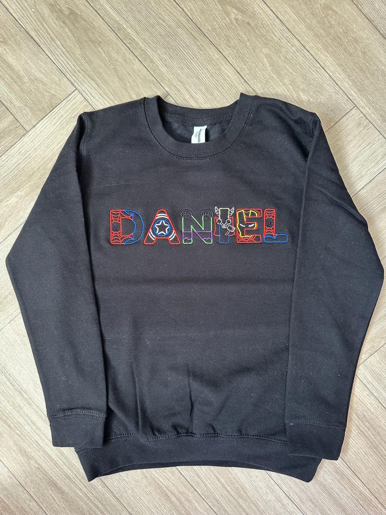Puede incluir: Sudadera negra de cuello redondo con el nombre "DANIEL" bordado en el pecho con letras coloridas con tem&aacute;tica de superh&eacute;roes. Las letras est&aacute;n delineadas en un color contrastante. La sudadera tiene mangas largas y pu&ntilde;os y dobladillo acanalados.