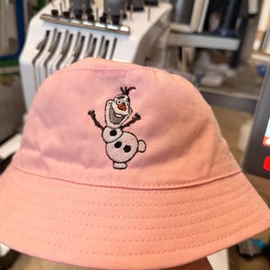 Puede incluir: Un sombrero de pescador rosa claro con un bordado de Olaf. El sombrero tiene un ala ancha y está hecho de una tela suave. El bordado de Olaf es en blanco, negro y naranja.