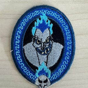 Peut inclure: Écusson brodé représentant Hadès, personnage du dessin animé Hercule de Disney. Le patch a une bordure ovale bleue et noire avec un motif détaillé. Hadès est représenté avec des flammes bleues pour les cheveux et un crâne en bas.