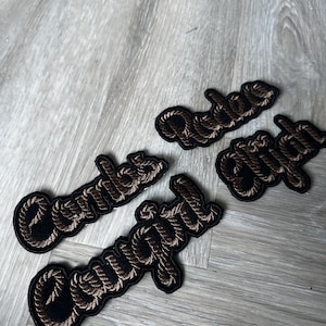 May include: Four embroidered patches with the words "Cowgirl" and "Rodeo" in a rope-like font. The patches are black with brown stitching, set against a light grey wooden surface.